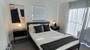A modern hotel room with a large bed, two bedside tables, a nightstand, a wall-mounted lamp, a window with blinds, and a curtain.