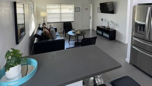 A modern kitchen with a central island, dining area with chairs, and a flat-screen TV mounted on the wall.