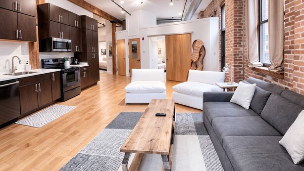 Una cocina moderna con armarios oscuros y electrodomésticos de acero inoxidable, una mesa de comedor de madera y un sofá gris en un salón luminoso con pared de ladrillo y ventanas amplias.