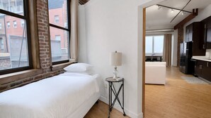 Un dormitorio con cama, una mesita de noche con lámpara, una ventana que da a vistas de edificios, y una cocina visible a través de un umbral abierto.