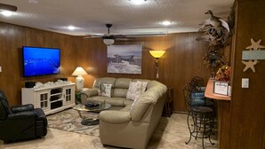 A living room with a beige sofa, a black leather chair, a glass coffee table, a white entertainment unit, a flat-screen TV, and a ceiling fan.
