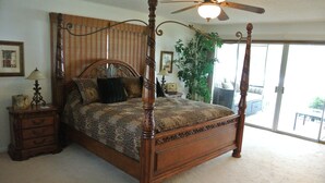 A four-poster bed with a canopy, a wooden nightstand with drawers, a floor lamp, and a large potted plant.