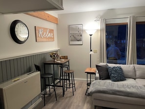 A cozy living room with a sofa, a dining table with chairs, a wall clock, a framed picture, and a floor lamp.