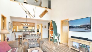 A spacious living area with a large TV, wooden flooring, and a kitchen in the background.