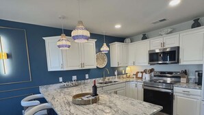 A modern kitchen with white cabinets, a marble island, bar stools, pendant lights, and stainless steel appliances.