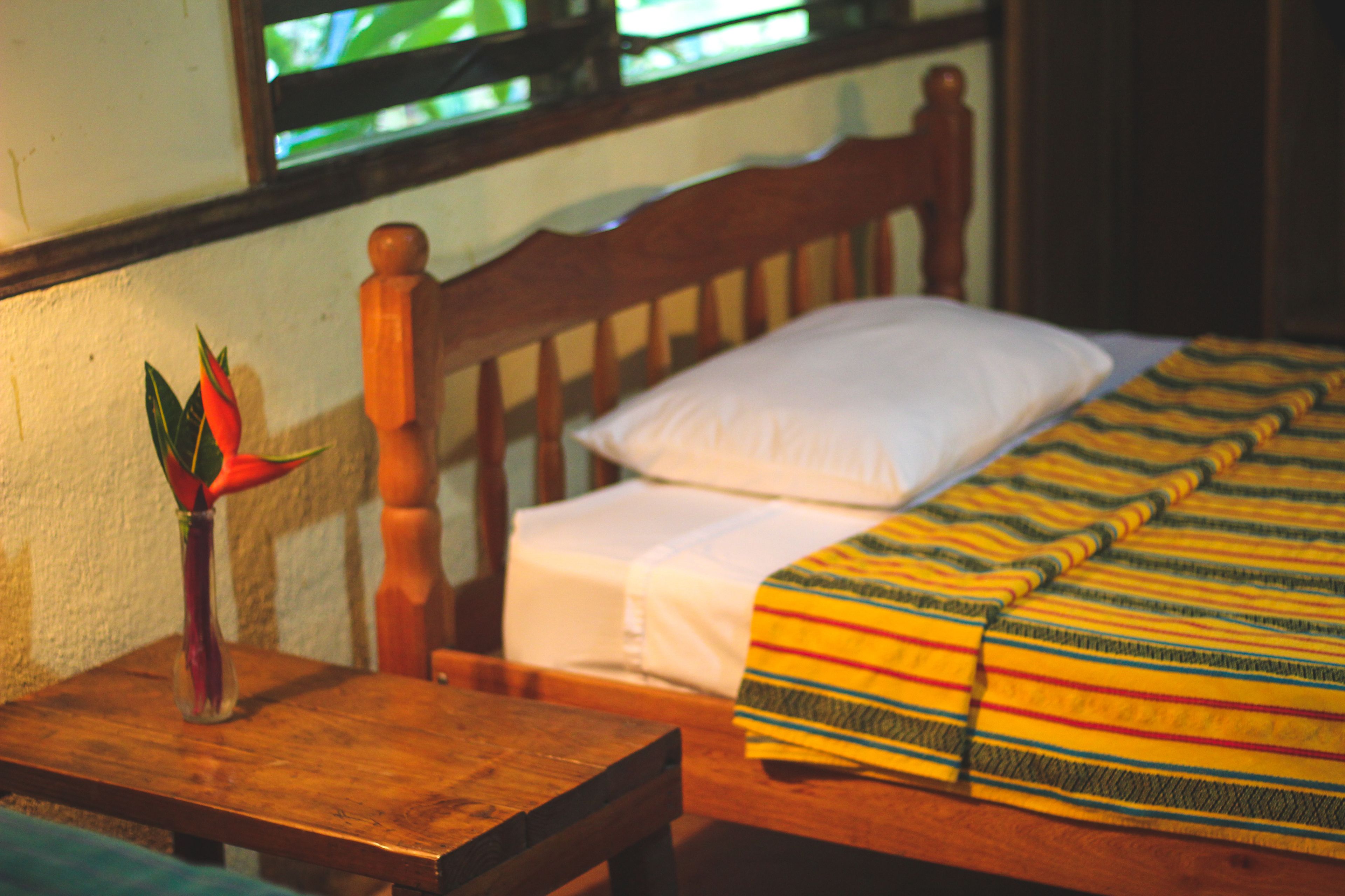 A wooden bed with a white pillow and a striped blanket, a wooden nightstand with a vase of flowers, and a window with a view of greenery.