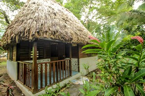 A thatched-roof hut with a wooden porch and a tiled floor, surrounded by lush greenery and a pink flowering plant. - Cohune Palms River Cabanas (San Ignacio)