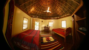 A room with a thatched roof, two beds with red and patterned blankets, a wooden chair, a small table with a lamp, and a window with blinds. - Cohune Palms River Cabanas (San Ignacio)