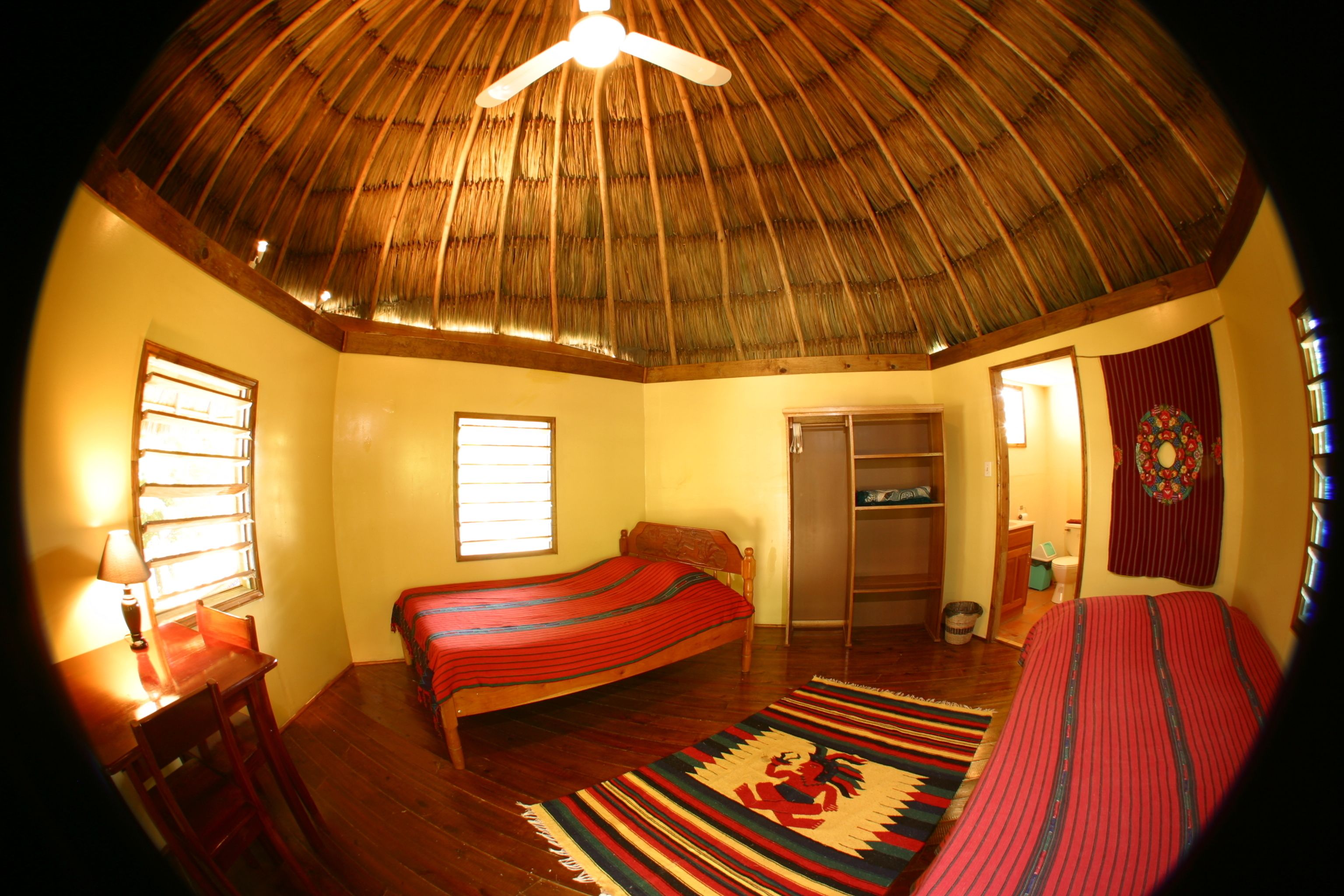 Una habitación con techo de paja, una cama con colcha a rayas rojas y azules, un escritorio de madera con lámpara, y una alfombra tejida y colorida.