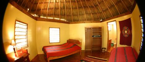 Una habitación con techo de paja, una cama con colcha a rayas rojas y azules, un escritorio de madera con lámpara, y una alfombra tejida y colorida.