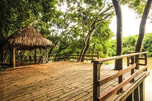 A wooden deck with a thatched-roof structure, surrounded by lush greenery and trees. - Cohune Palms River Cabanas (San Ignacio)