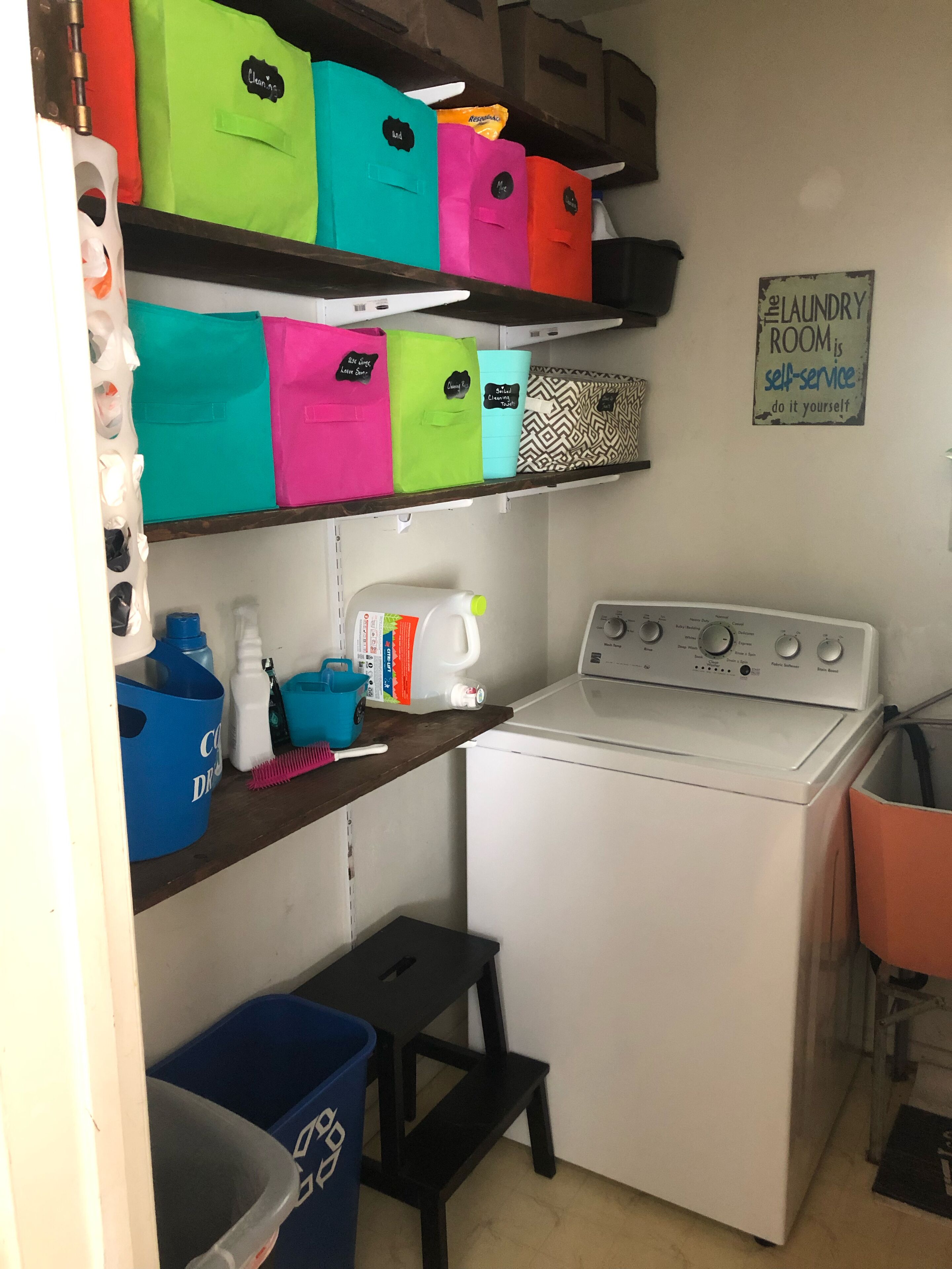 Une buanderie équipée d’un lave-linge, d’étagères avec des bacs de rangement colorés et d’un petit tabouret.