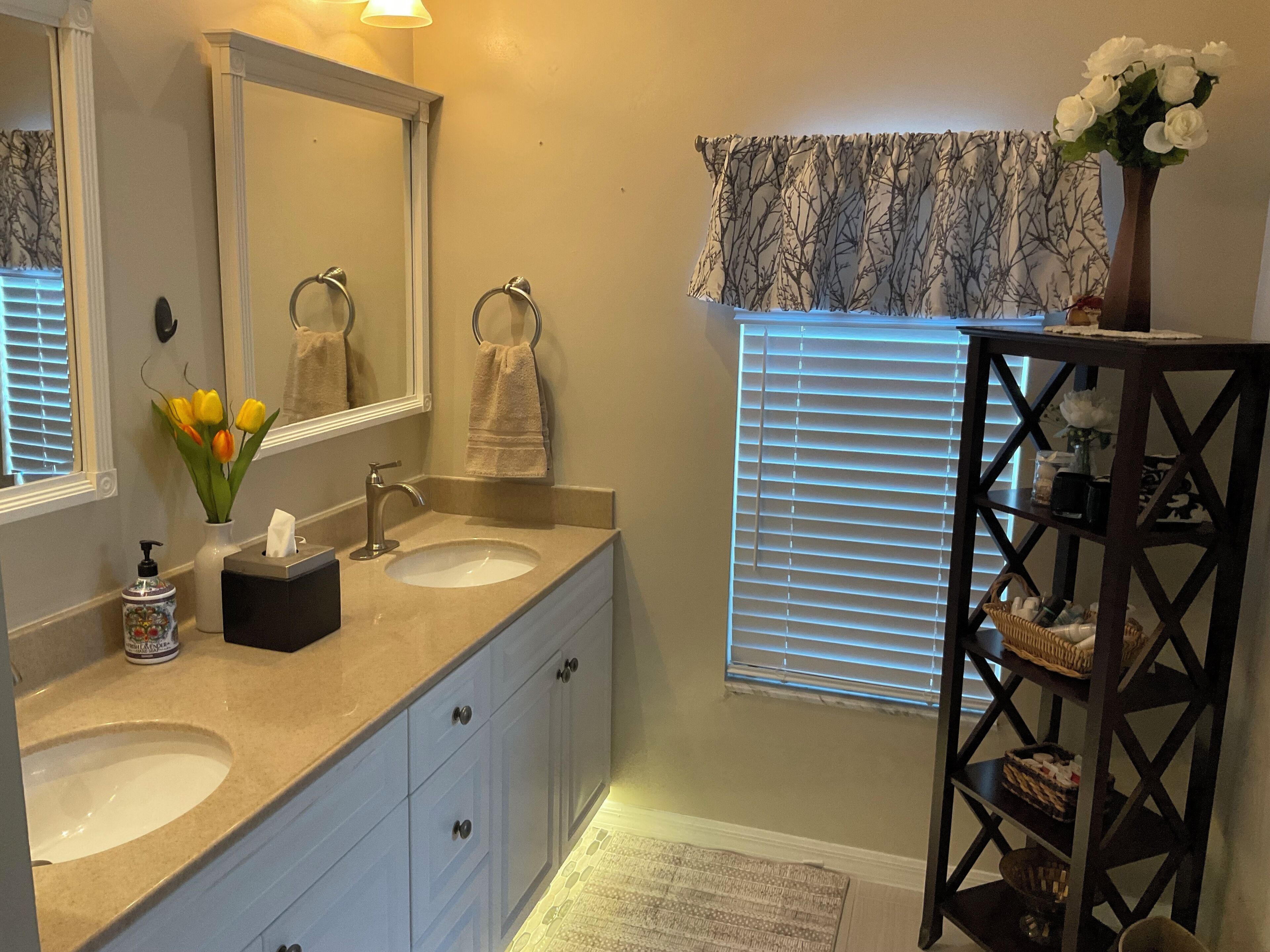 A bathroom with two sinks, a mirror, a towel rack, a vase with flowers, and a window with blinds.