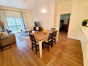 Uma sala de jantar com mesa e cadeiras de madeira, televisão em um suporte e varanda com vista.