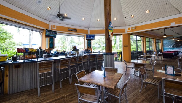 Un restaurant avec un plafond haut, des tables et des chaises en bois, un coin bar et de grandes fenêtres donnant sur l’extérieur.