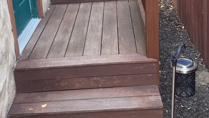 Une terrasse en bois avec des marches menant à une porte, un grand bac à fleurs et une petite statue.