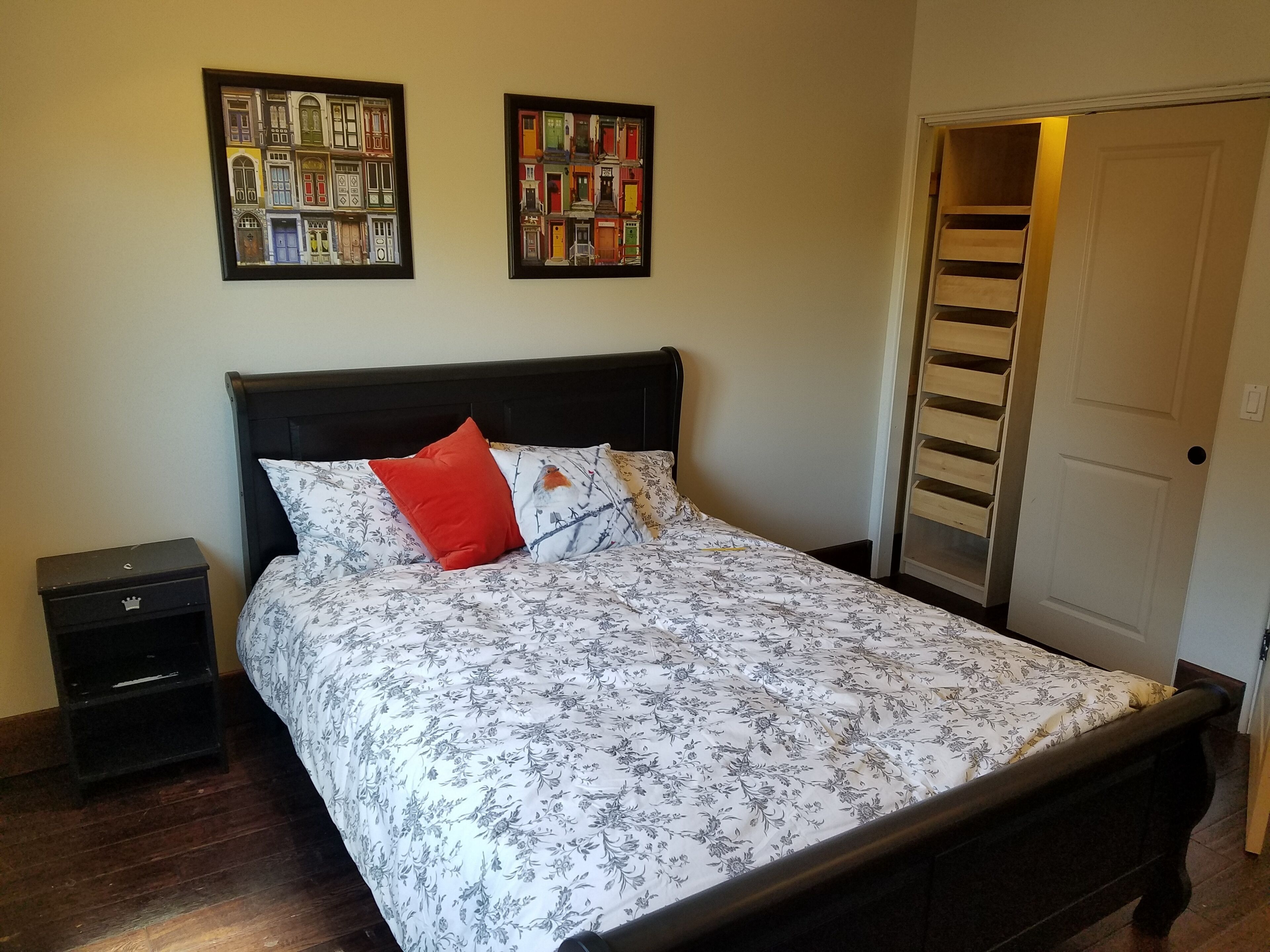 Una cama bien hecha con ropa de cama floral, una mesita de noche con cajón y dos cuadros en la pared.