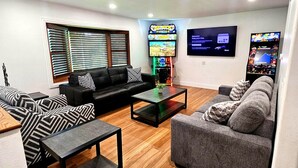 A modern living room with a black leather sofa, grey sofa, and a coffee table. There are two flat-screen TVs mounted on the wall, one displaying a game and the other a welcome screen. - All Decked Out | Arcade Games | Amazing 5 BR |  Huge Deck | Fire Pit | Close to Everything WI Dells (Wisconsin Dells)