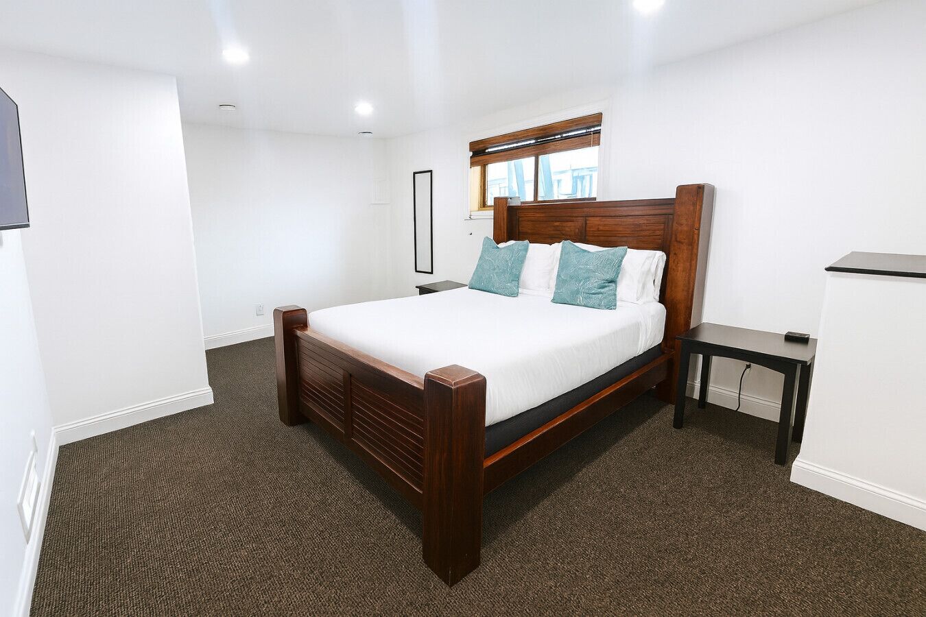 A bedroom with a wooden bed, a small bedside table, a mirror, and a window with blinds.