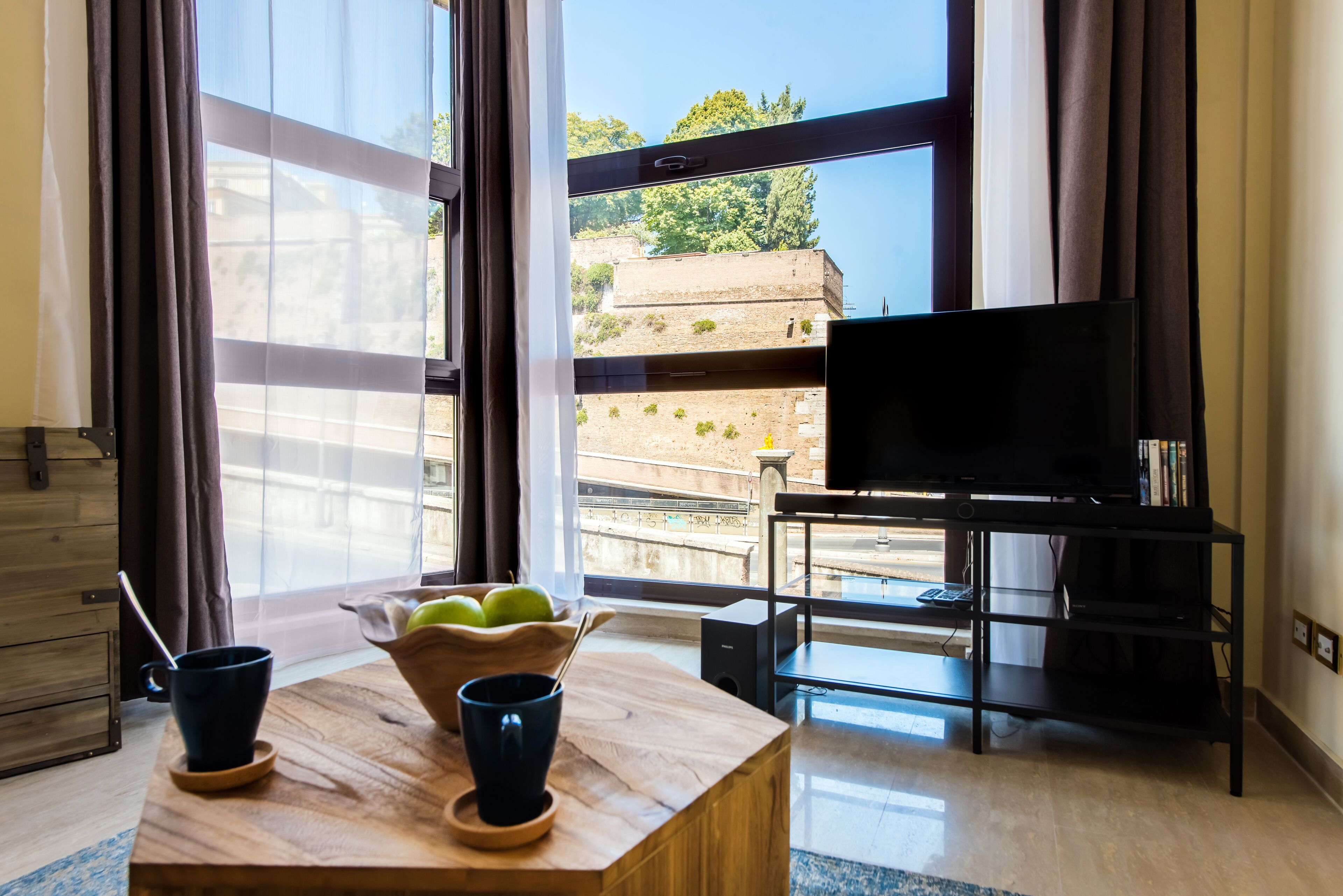 A room with a wooden table, two cups, a bowl of apples, a TV on a stand, and large windows with curtains.
