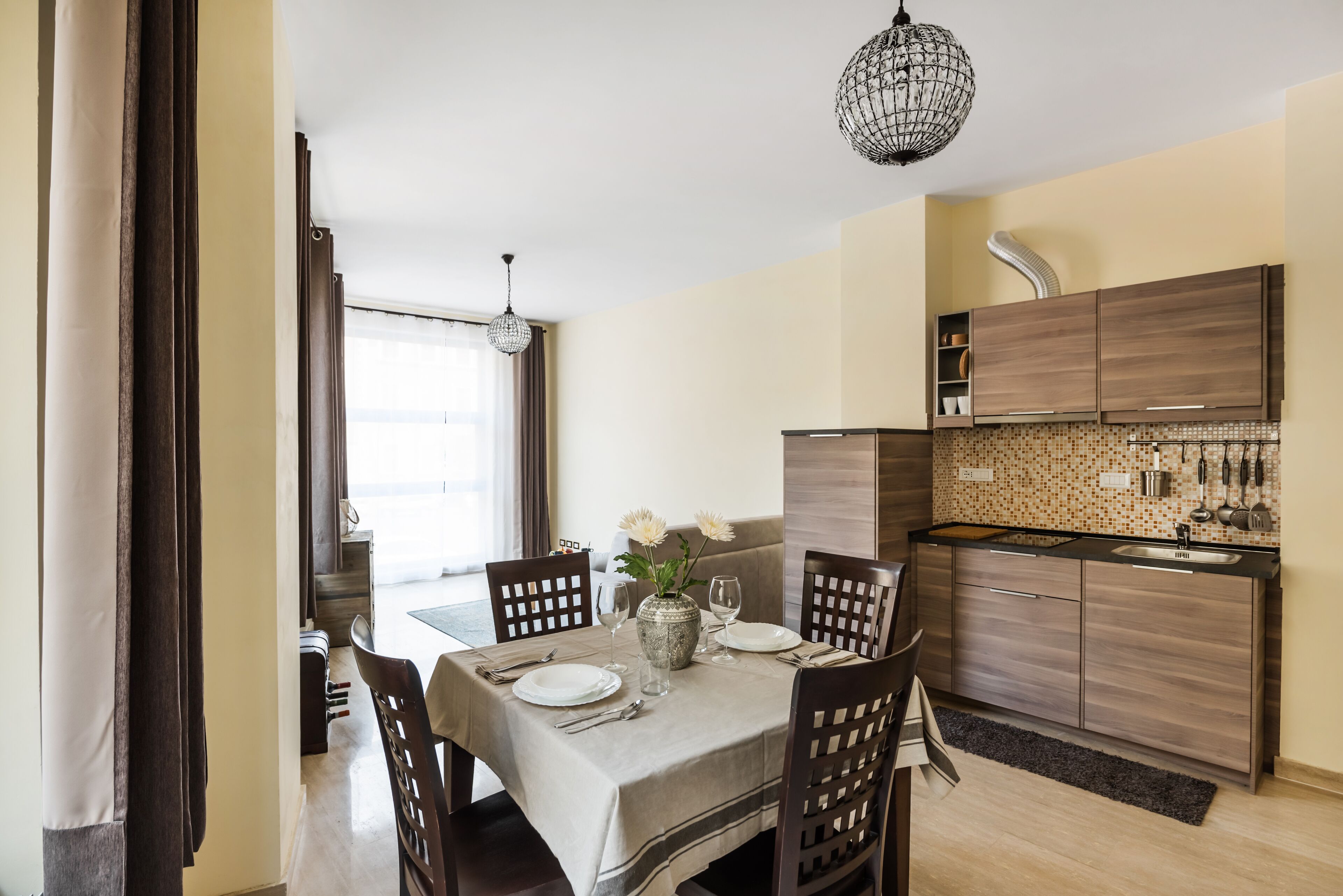 A dining area with a table set for four, a vase with flowers, and a kitchen in the background.