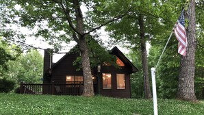 En rustik hytte med veranda, omgivet af træer og et flagstang med det amerikanske flag.