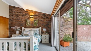 A bedroom with a brick wall, a bed with a white frame, a nightstand, a dresser, and a window with a view of the outdoors. - Kings Point Lake House - Family Getaway
Wake up to peaceful lake views (Kings Point)