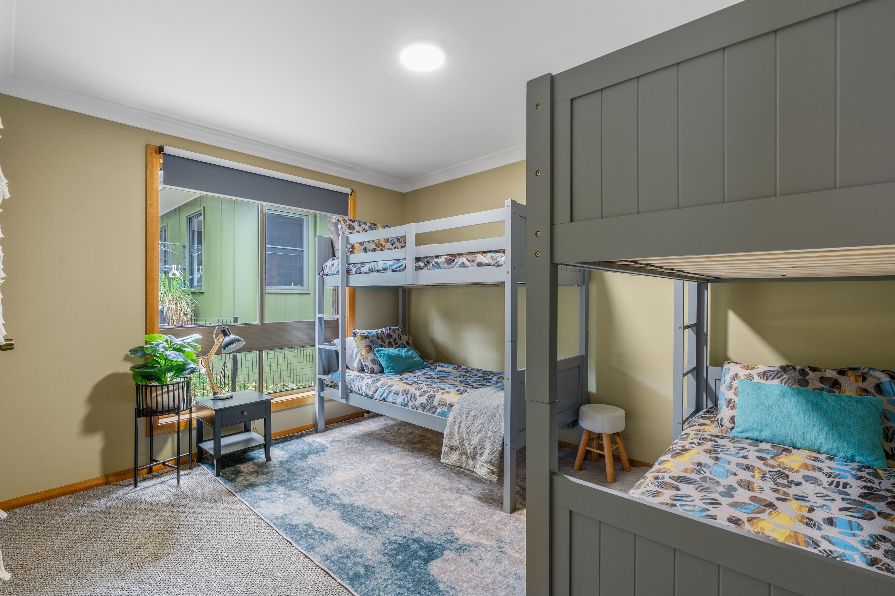 Een slaapkamer met stapelbed, bureau en stoel, een klein krukje en een potplant.