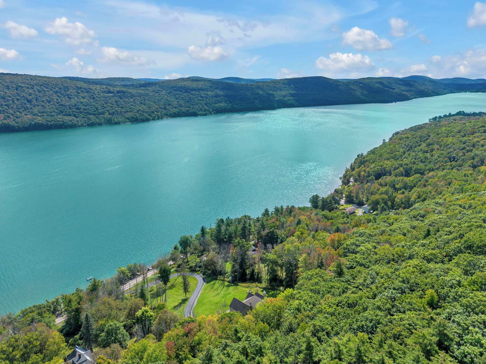 Ein See, umgeben von bewaldeten Hügeln, mit einer Straße und Gebäuden im Hintergrund.