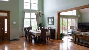 A dining area with a wooden table and chairs, a chandelier, and large windows allowing natural light. - Grand Slam - Lake House Cooperstown Dreams Park (Cooperstown)