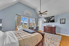 Une chambre à coucher comprenant un lit, une commode, un ventilateur de plafond, une grande fenêtre avec des rideaux et un téléviseur fixé au mur.
