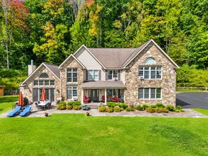 Une maison en pierre à deux étages, dotée d’un porche à l’avant, d’un patio couvert avec des meubles d’extérieur et d’une pelouse bien entretenue.