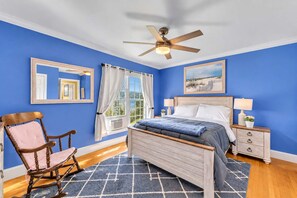 A bedroom with a bed, a chair, a nightstand, a ceiling fan, and a window with curtains.