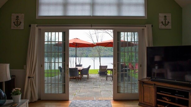 A room with a large TV, a dining area with outdoor umbrellas, and a view of a lake through glass doors. - Grand Slam - Lake House Cooperstown Dreams Park (Cooperstown)