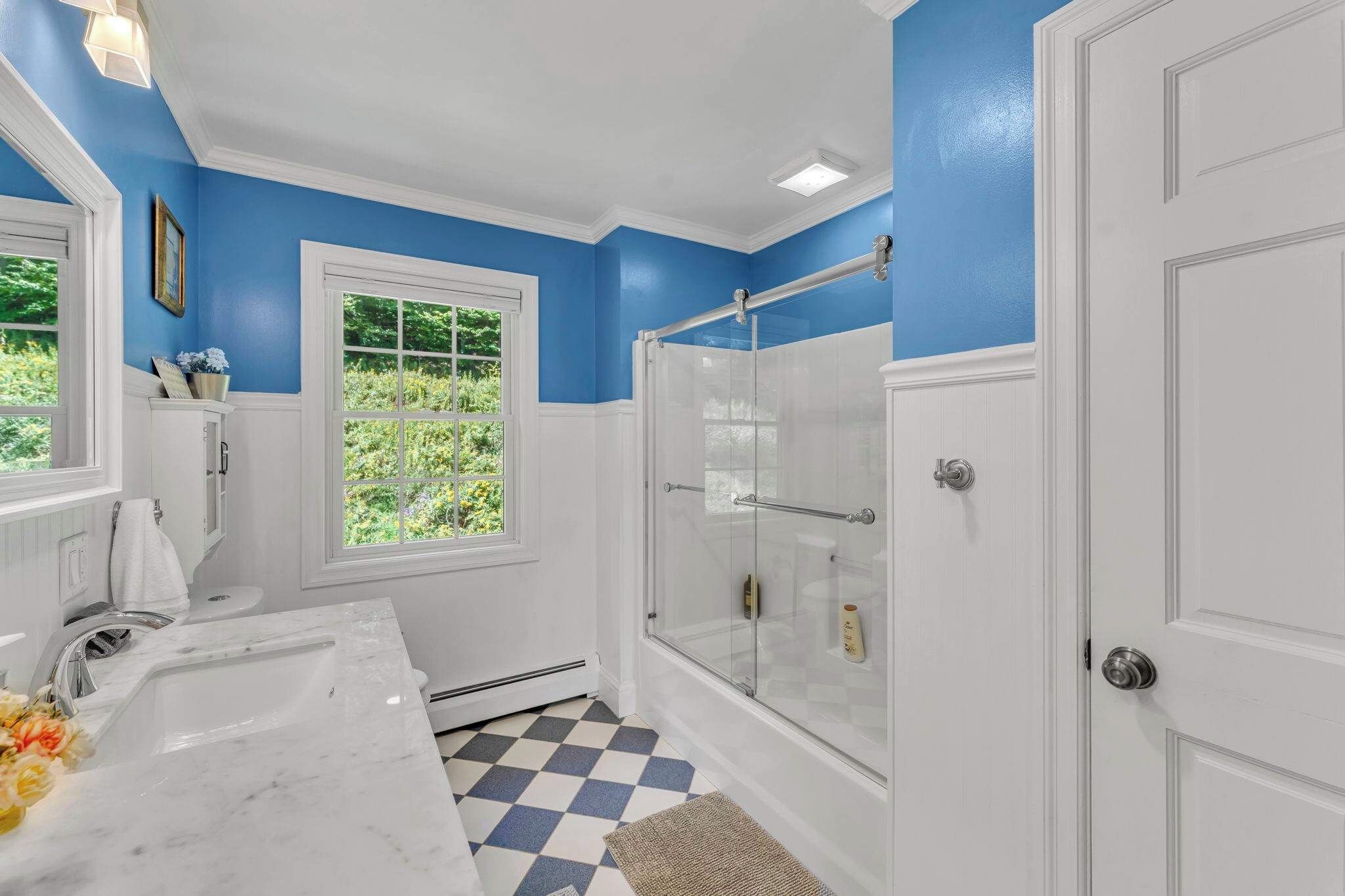 Une salle de bain avec une grande baignoire blanche, une douche avec des portes en verre, une fenêtre donnant sur un paysage verdoyant et un carrelage à motifs carrés.
