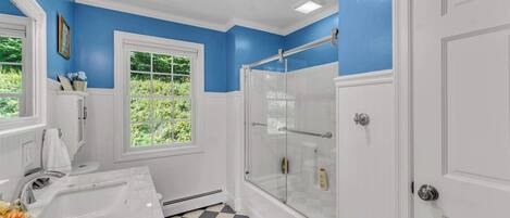 Une salle de bain avec une grande baignoire blanche, une douche avec des portes en verre, une fenêtre donnant sur un paysage verdoyant et un carrelage à motifs carrés.