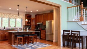 A spacious kitchen with wooden cabinets, a large island with bar stools, stainless steel appliances, and a view of a staircase. - Grand Slam - Lake House Cooperstown Dreams Park (Cooperstown)