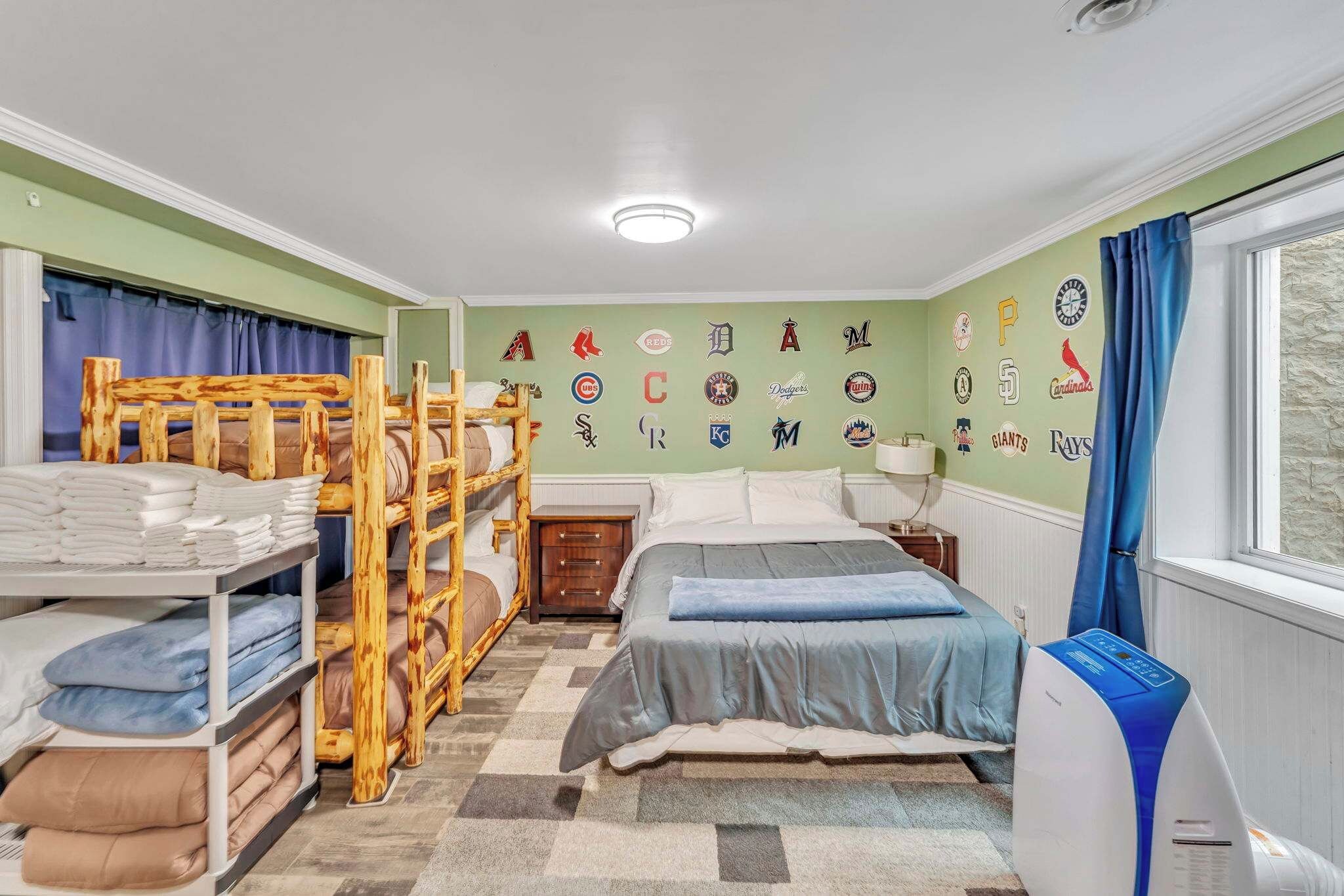 A bedroom with bunk beds, a bed, a nightstand, a lamp, a heater, and a wall with sports team logos.
