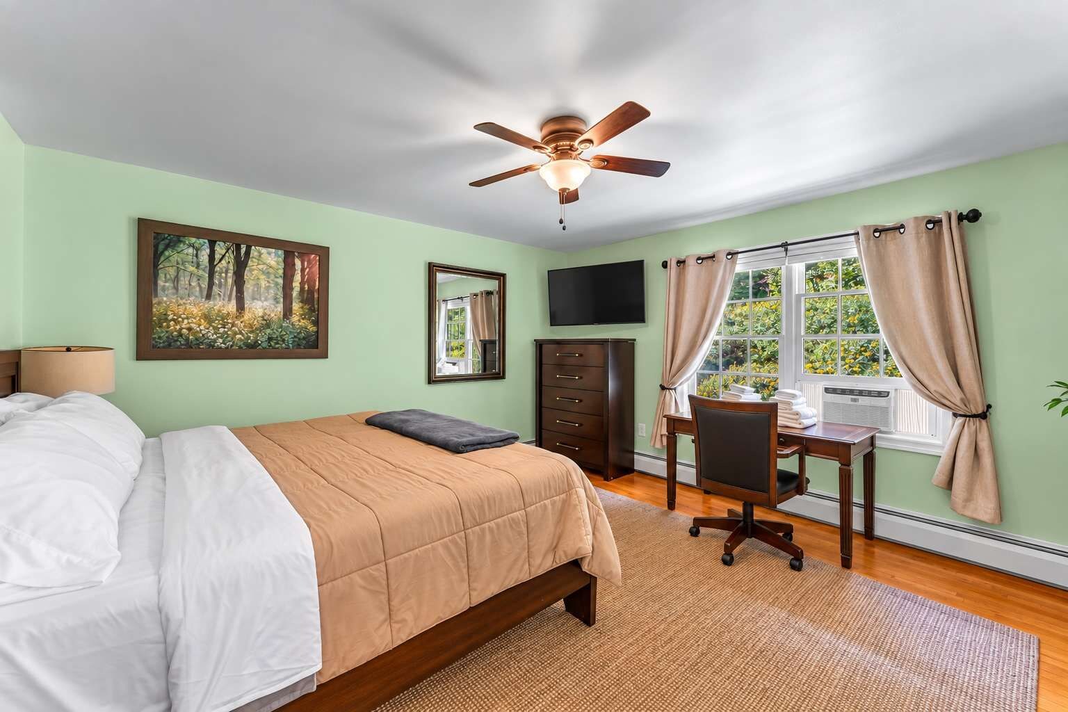 Une chambre à coucher comprenant un lit, un bureau, une chaise, un ventilateur de plafond, une fenêtre avec des rideaux, un miroir et un téléviseur.