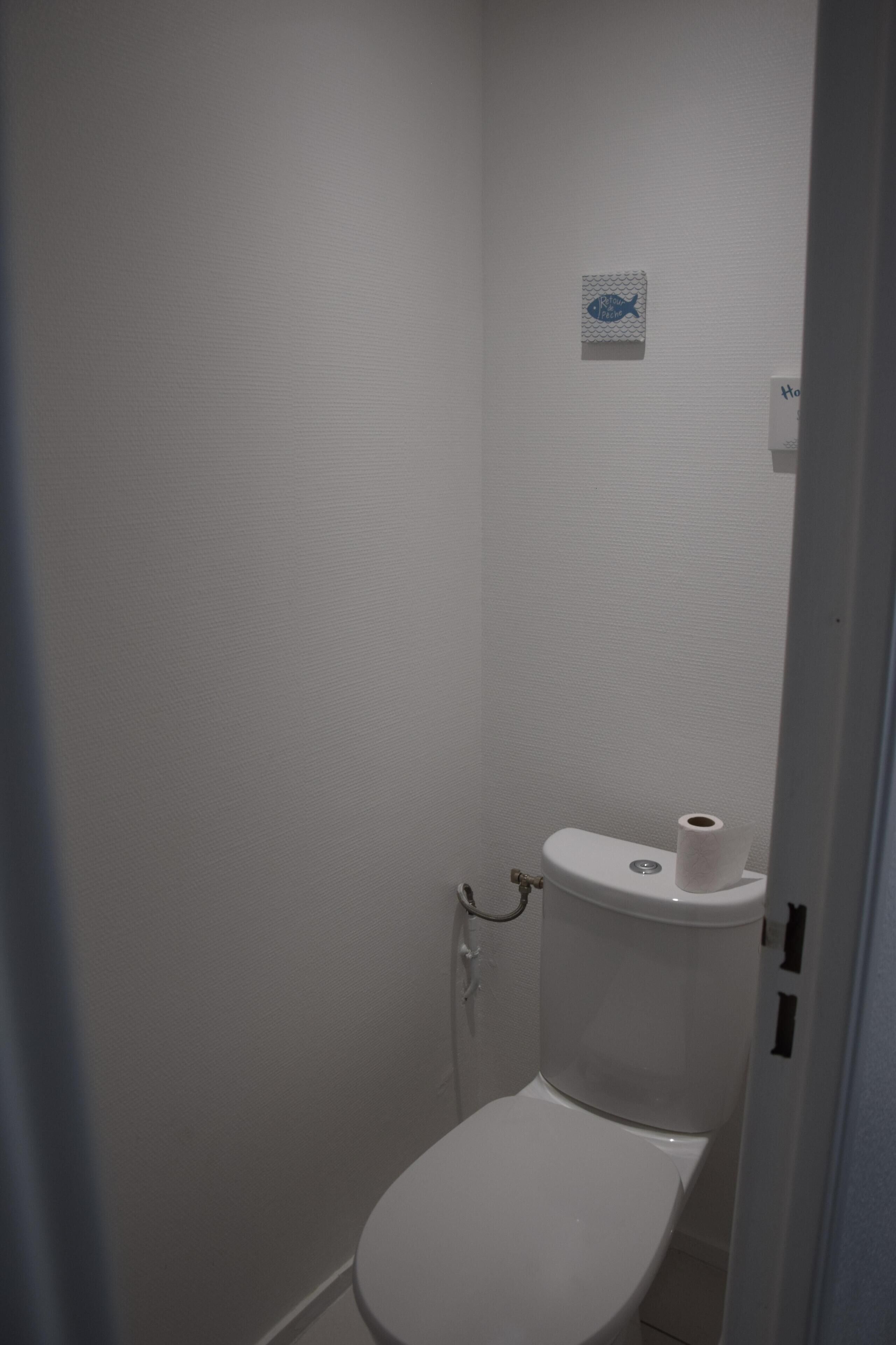 A small, white, single-occupancy bathroom with a toilet, a paper towel holder, and a small wall-mounted sign.