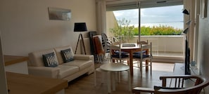A living room with a sofa, a table, chairs, a lamp, and a view of the sea through a large window.