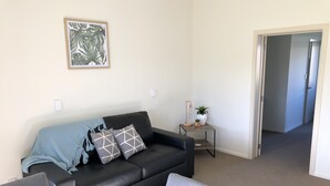 Una sala de estar moderna con un sofá de cuero negro, una mesa de centro y un sillón gris. Hay una mesita auxiliar con una lámpara y una planta en maceta. Un cuadro enmarcado cuelga en la pared, y una puerta da a otra habitación.