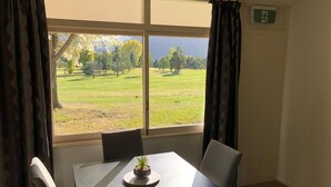 Una habitación con una mesa y sillas, una ventana que ofrece vistas a un campo verde y árboles, y una pequeña planta en maceta sobre la mesa.