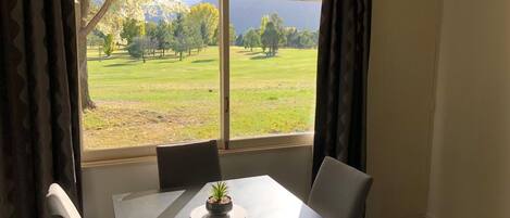 Una habitación con una mesa y sillas, una ventana que ofrece vistas a un campo verde y árboles, y una pequeña planta en maceta sobre la mesa.