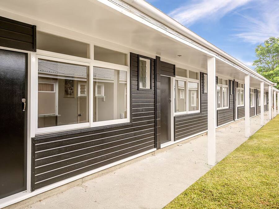 A row of motel rooms with exterior sliding doors and windows, a covered walkway, and a grassy area.
