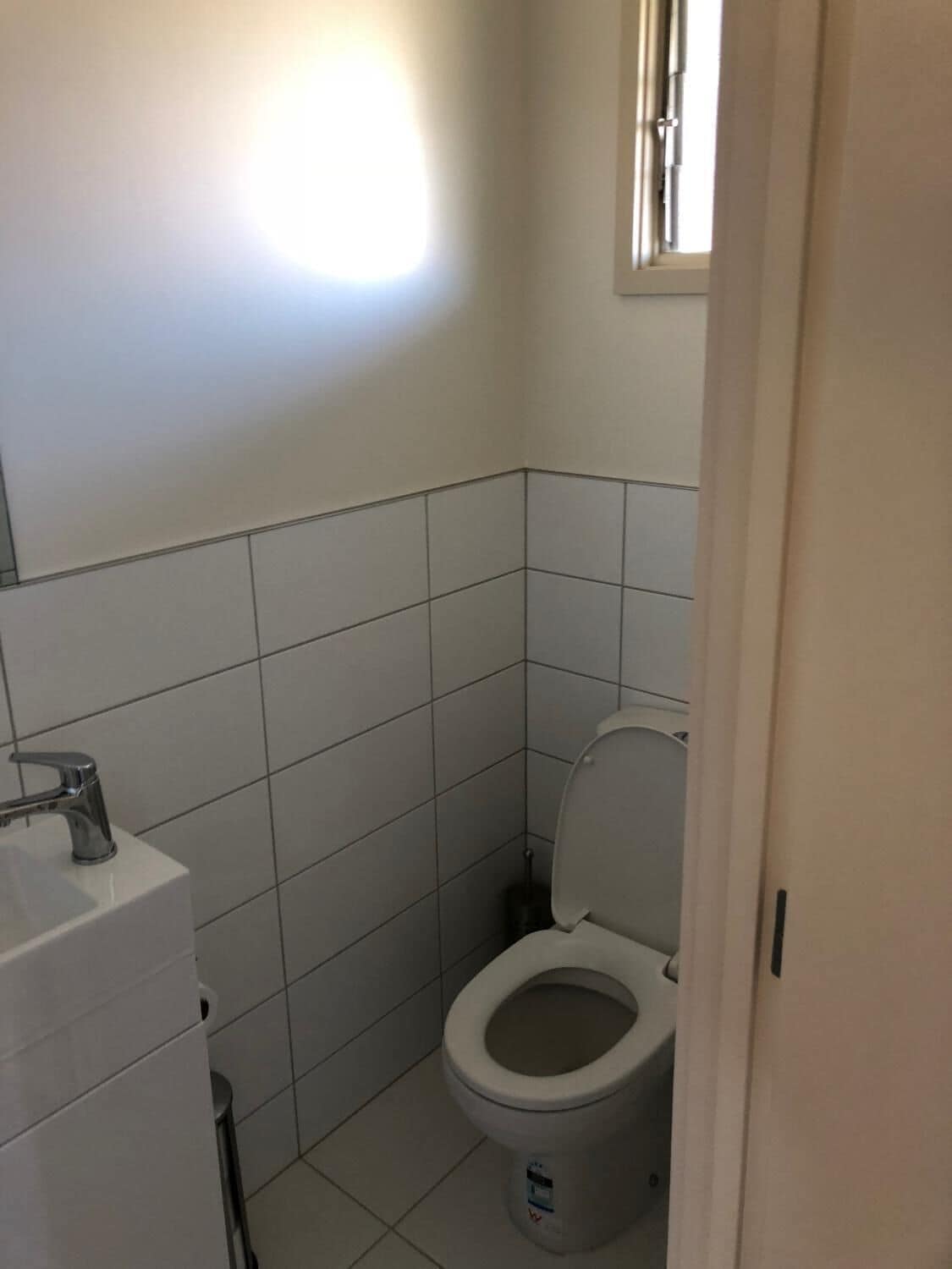Ein kleines, sauberes Badezimmer mit weißen Fliesen, einer Toilette, einem Waschbecken und einem Fenster.
