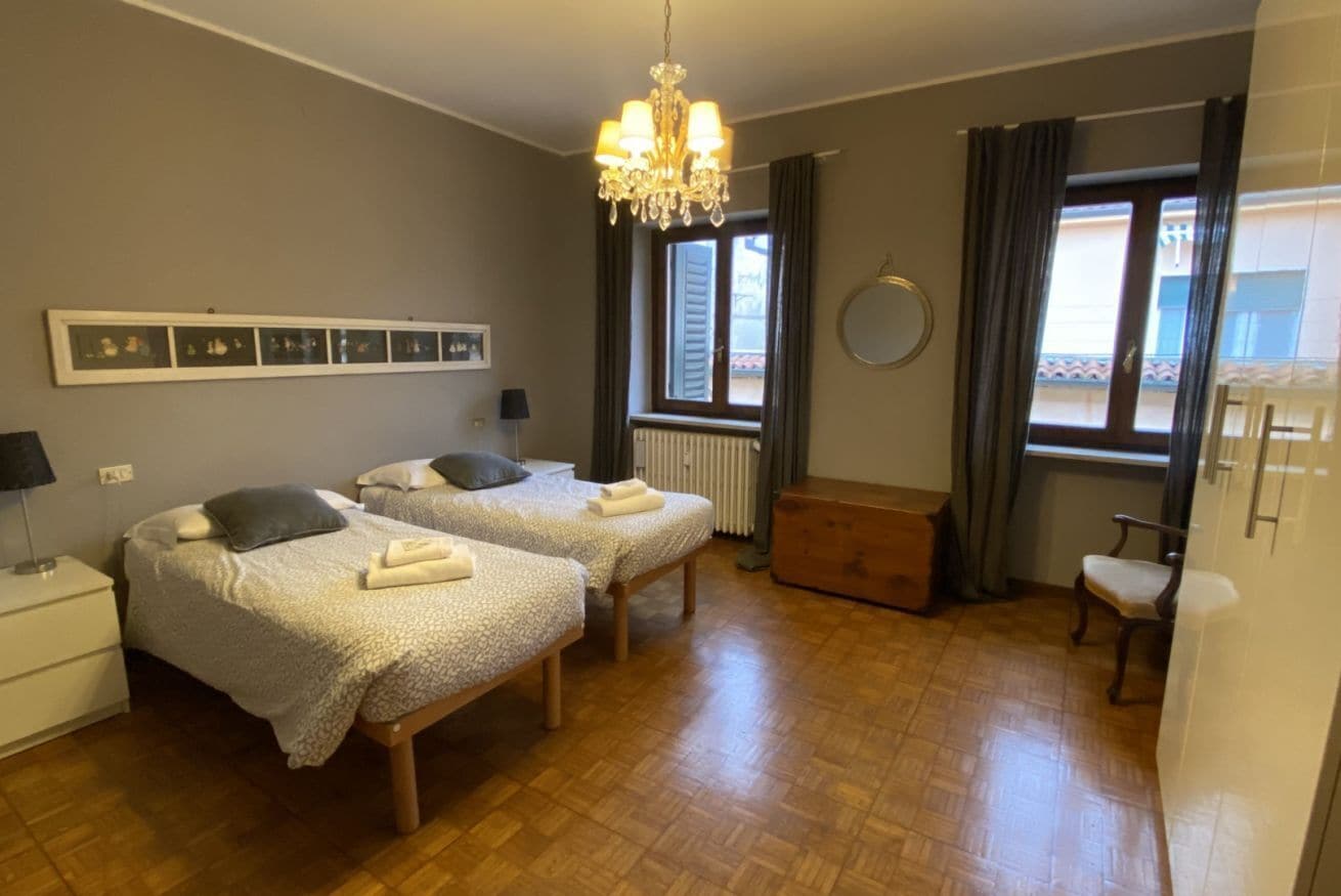 A room with two beds, a wooden bench, a radiator, a window with curtains, a chandelier, and a wall-mounted mirror.