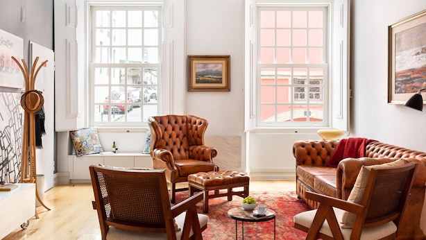 Un salon chaleureux avec un canapé en cuir marron, une table basse en bois et un fauteuil en osier. Il y a deux fenêtres à cadres blancs, un tableau au mur et un porte-chapeau avec un chapeau.
