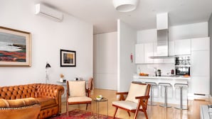 Un salon moderne avec un canapé en cuir, deux fauteuils, une petite table et un coin cuisine doté de meubles blancs et d’une hotte de cuisine en acier inoxydable.