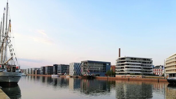 Ein Hafen mit einem großen Segelschiff, modernen Gebäuden und ruhigem Wasser, das die Architektur spiegelt.